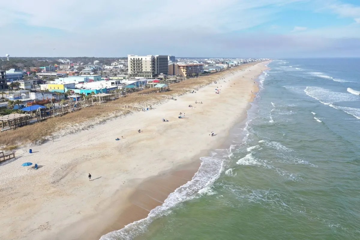 A wide perspective of the beach, boardwalk, and adjacent town, showing people walking, shopping, and engaging in water sports. The scene includes sunset lighting and hints of festival activity, underlining the area's appeal for both day and evening exploration.