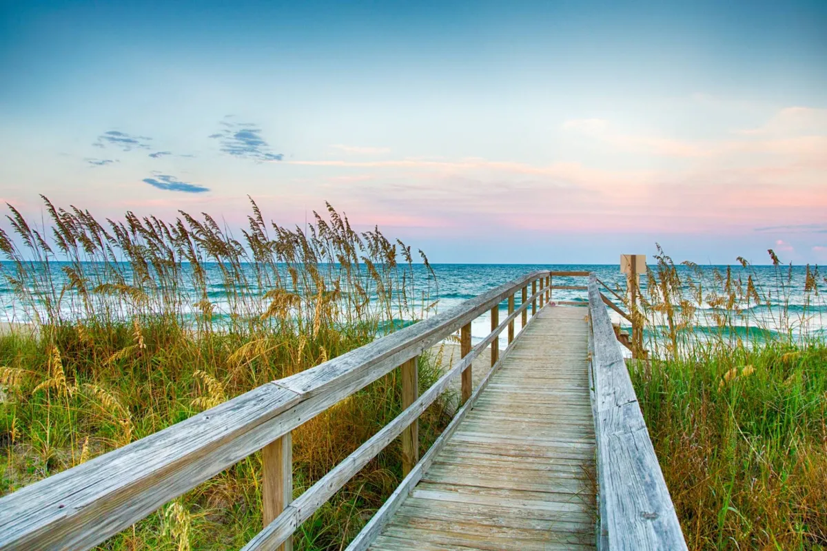 A serene, panoramic view down a boardwalk framed by dune grasses, opening onto the expansive ocean and sky. The scene captures the magic of walking to the shore, with ample space for activities like frisbee, sandcastle building, or sunset watching.
