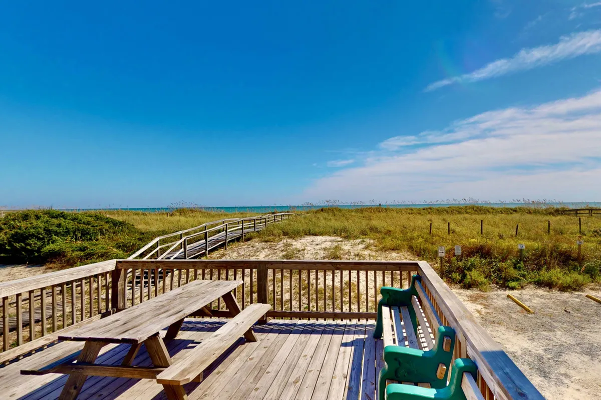 The image showcases a classic wooden deck with a picnic table and green benches overlooking the ocean. A private boardwalk leads directly from the house to the sandy beach, with unobstructed shoreline views perfect for watching sunsets or sipping morning coffee. The scene exudes a relaxed, nostalgic beach cottage vibe, inviting guests to unwind and reconnect with simple pleasures.