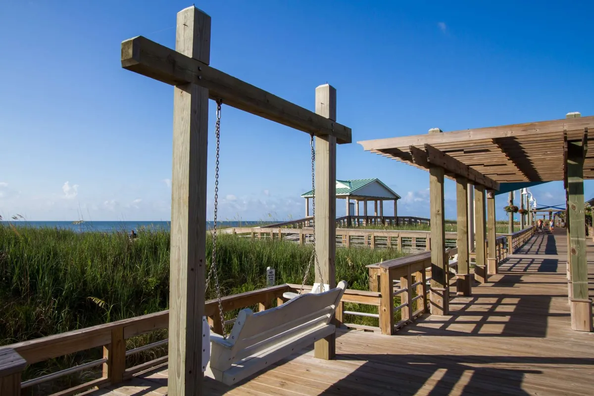 A tranquil outdoor scene featuring a wooden swing set along a boardwalk, with pavilions in the background and lush greenery all around. The view hints at a relaxing environment ideal for unwinding and sharing stories, with ocean breezes indicated by the open layout.