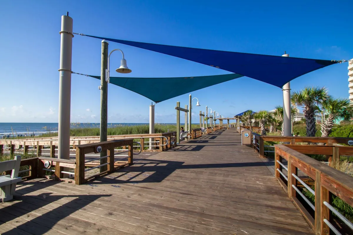 A vibrant stretch of the Carolina Beach boardwalk, complete with colorful shade sails, palm trees, wooden paths, lampposts, and an ocean view in the distance. The area features welcoming seating and a lively, communal atmosphere, inviting visitors to stroll, relax, or gather for public events.