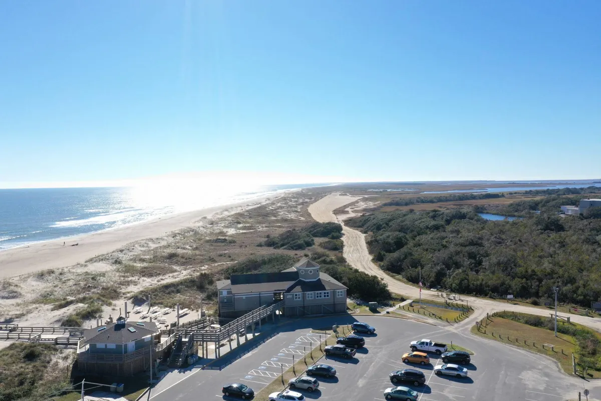 A sweeping view of the ocean, shoreline, parking area, and the beach house itself, set against a serene backdrop. The image showcases the proximity to beach activities and the expansive coastal environment accessible from the property.
