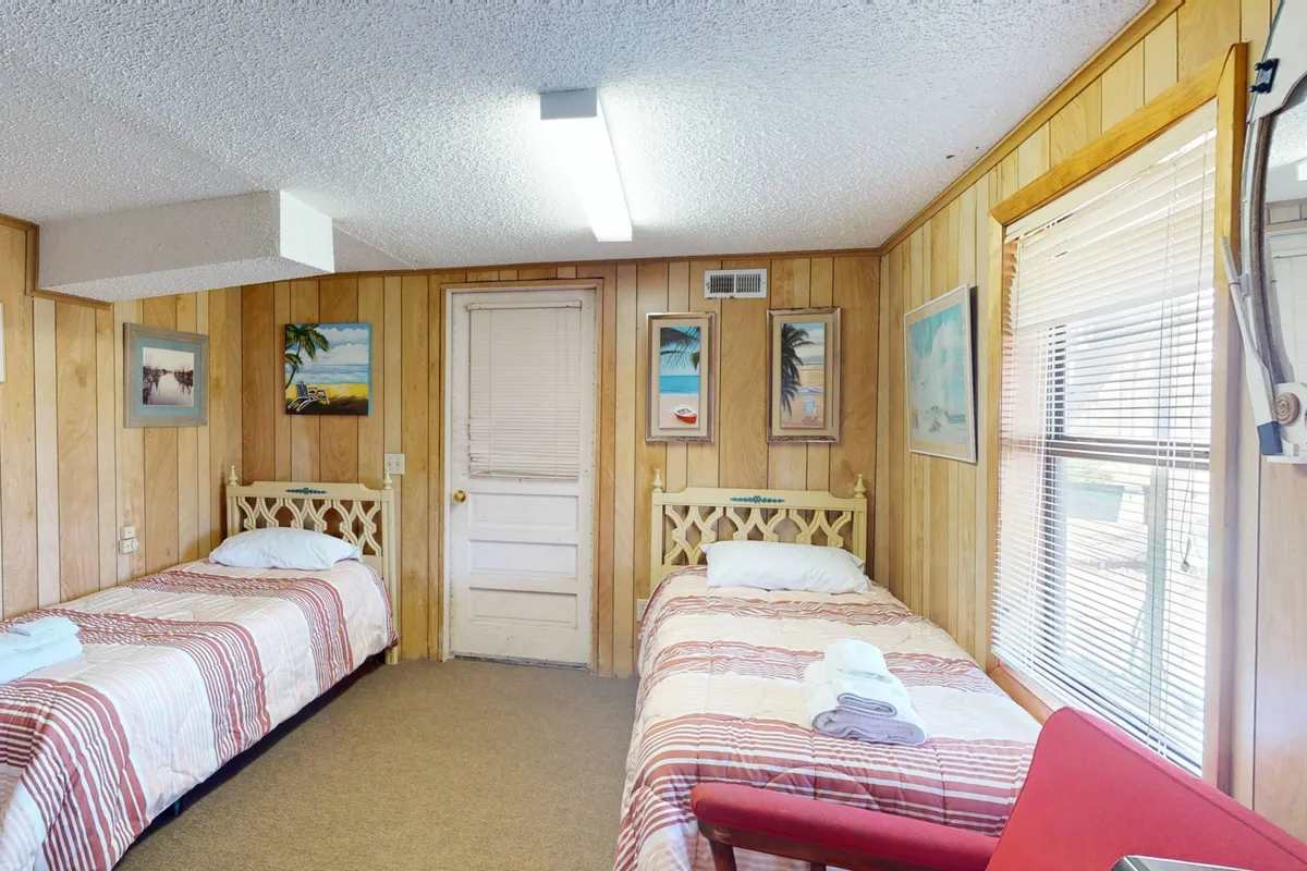 A homey bedroom with two twin beds covered in striped linens, wooden wall paneling, and a door leading outside. Decorative paintings and a window with blinds add personal touches, while the exterior door hints at a private entrance.