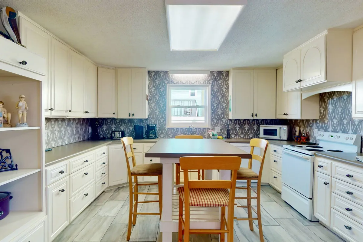 A bright, welcoming kitchen with a central island and chairs, decorative wall pattern, and sunlight streaming through a window. The space is open and inviting, designed for casual dining, cooking, and gathering with family or friends.