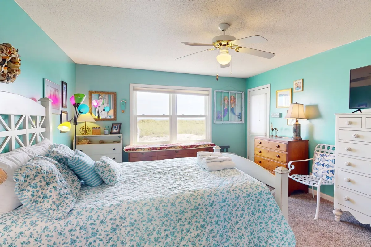 A sun-filled bedroom featuring a bed with cheerful floral bedding, a colorful floor lamp, and decorative wall art. A large window lets in natural light, giving the room a bright and airy feeling while vintage touches like the dresser and artwork evoke classic beach cottage style.