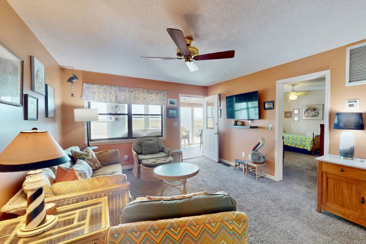 A relaxed, open-plan living and dining area with plush seating, large windows framing unobstructed ocean views, and charming decorative wall art. The arrangement invites casual conversation, reading, or enjoying movie nights as the sun sets over the waves.