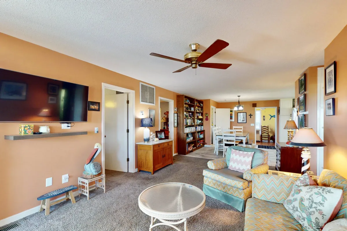 A bright, inviting living area featuring a comfortable seating arrangement with plush sofas and armchairs, a decorative bookshelf filled with books, and ambient light fixtures. The windows let in ample natural light, highlighting the warm tones of the original hardwood floors and vintage décor. The space feels open and perfect for reading, socializing, or family gatherings.