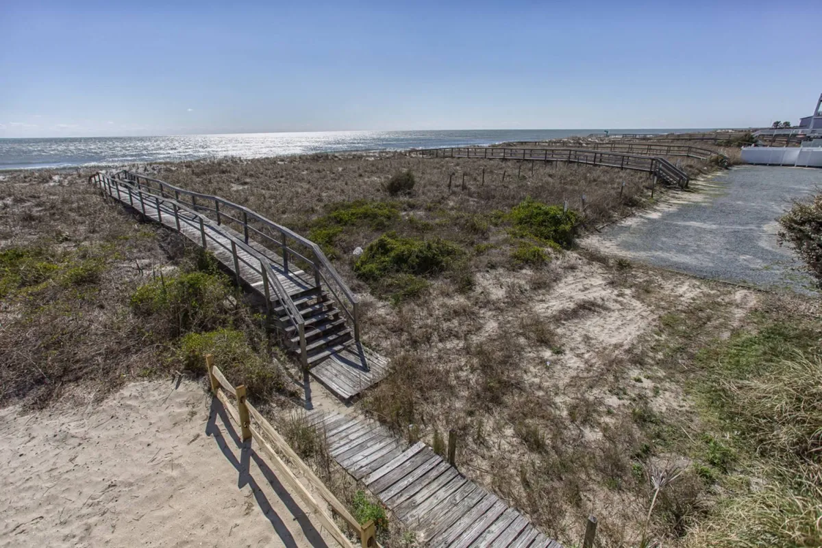 A serene wooden boardwalk stretches over sandy dunes toward the sea. The sun casts a warm glow, and gentle waves roll in, setting a peaceful scene ideal for leisurely walks or family outings. The image emphasizes the connection between the Boardwalk and the natural beauty of the beach.