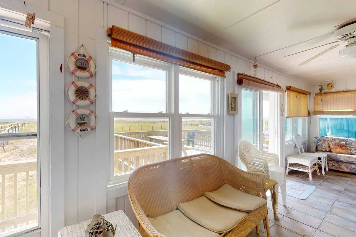 A tranquil sunroom featuring woven chairs, large windows with sweeping views of the ocean, and tasteful decorative accents. The setting is ideal for gazing out at the waves, sharing quiet stories, or simply relaxing in the sun-dappled space.