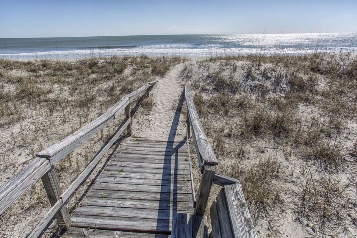 A wooden walkway leads directly from the property through a sandy path to the expansive beach and rolling waves. The perspective places viewers at the start of their private journey to the sand, with serene ocean views stretching out ahead.
