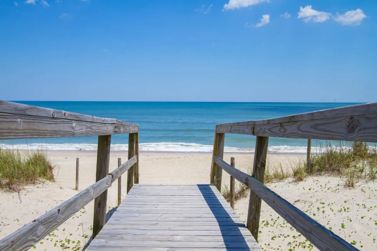 This exterior scene highlights a broad wooden pathway leading over the dunes to a sweeping sand beach, with the ocean horizon and blue sky beyond. The setting suggests opportunities for sunrise coffee, sunset toasts, and relaxed beach outings mere steps away.
