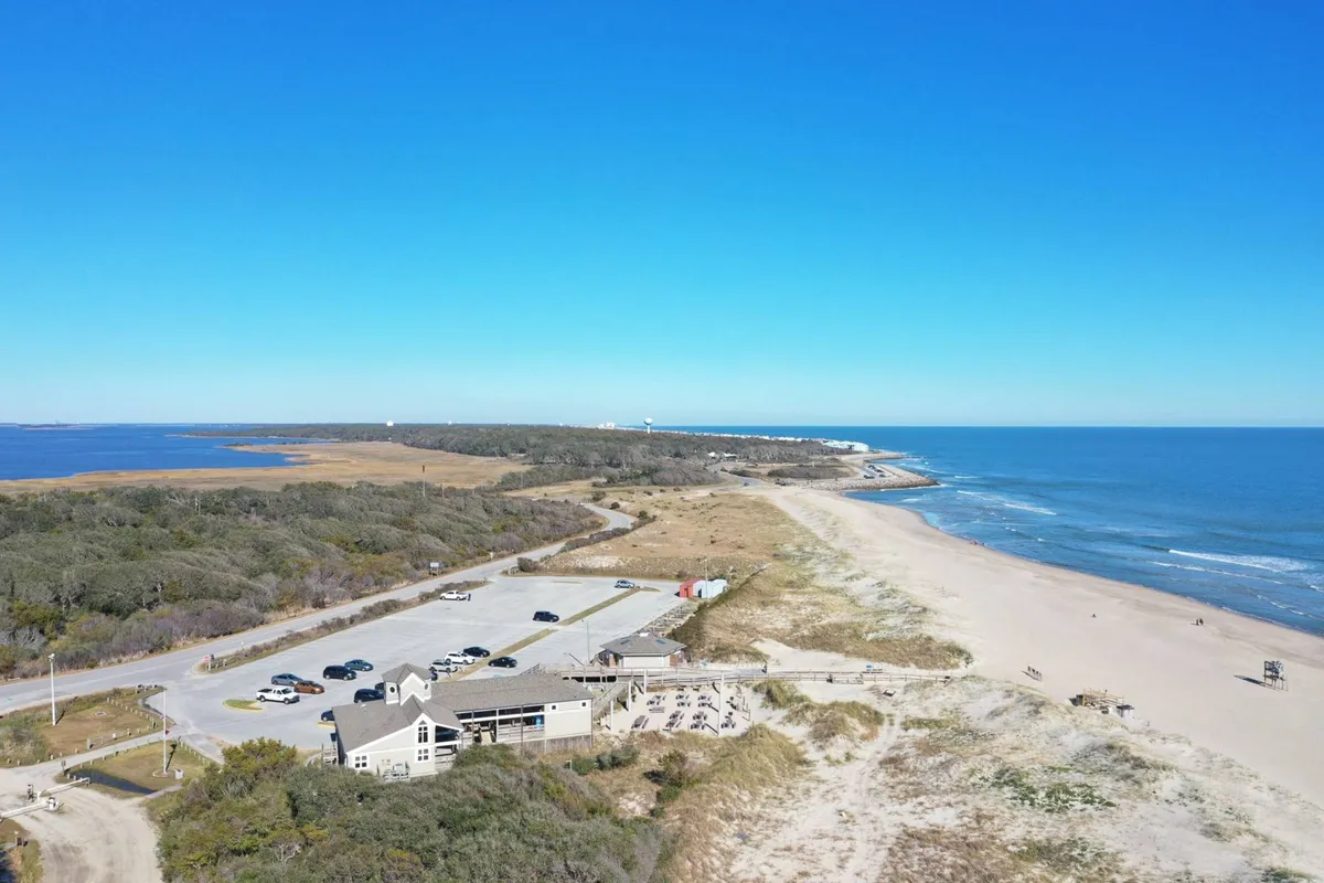 A vibrant view of the beach with the ocean in the background, a lively parking lot, a casual restaurant, and the sprawling coastal landscape. Beachgoers can be seen enjoying the sun while the nearby eatery hints at local dining options. The image captures both natural beauty and easy access to local flavors.