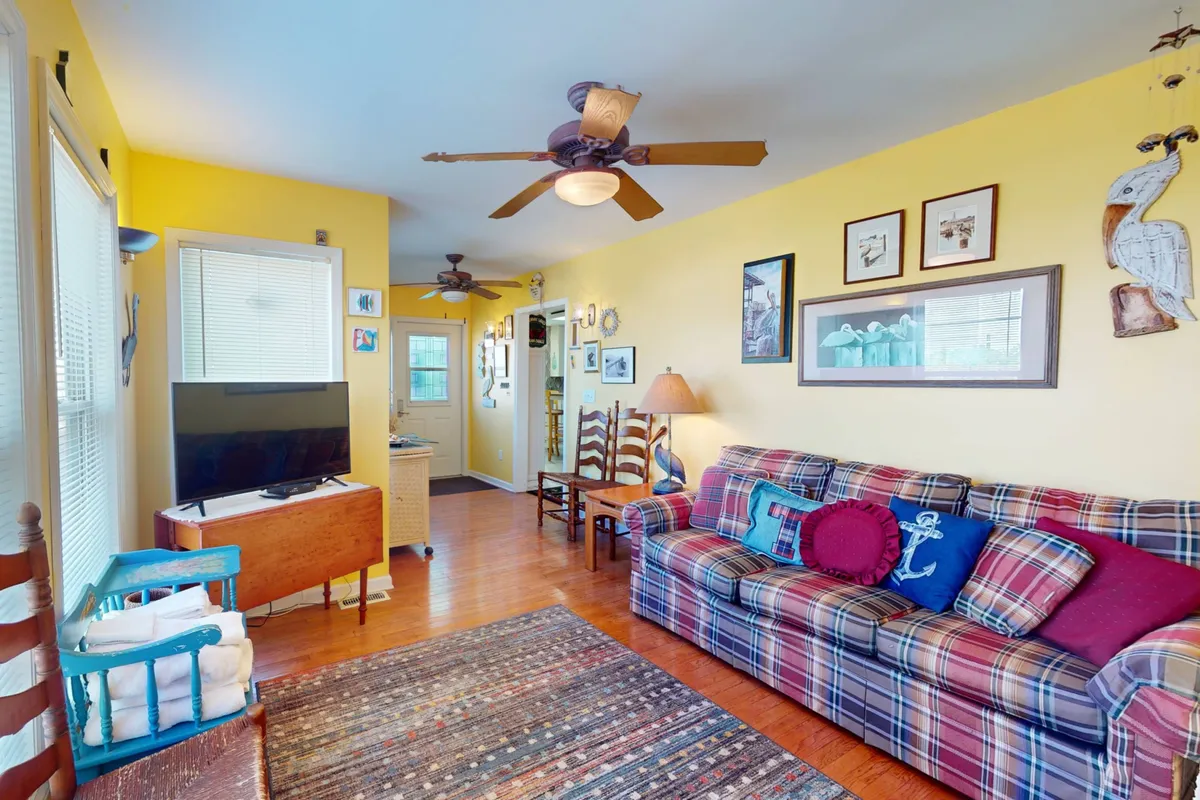 A cheerful living space with a plaid sofa, colorful throw pillows, wall art, wooden chairs, and a television. The room is designed for relaxing, socializing, watching TV, reading, or playing games, evoking a sense of comfort and togetherness.