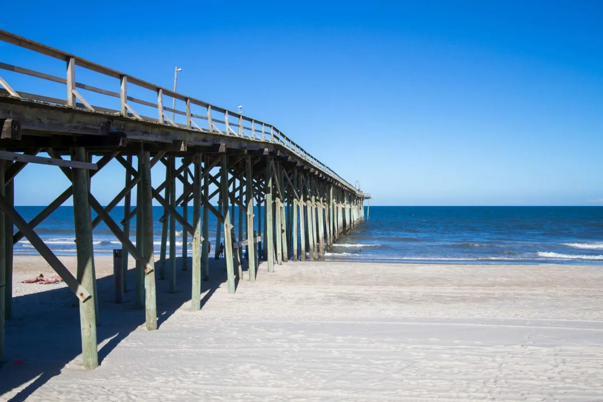 A classic wooden pier stretches out over the ocean with soft sand at its base, creating a quintessential coastal scene. The water glistens in the sunlight, and the quiet pier invites both fishing and leisurely strolls. The open sky and gentle waves embody the relaxed, natural beauty of Carolina Beach.