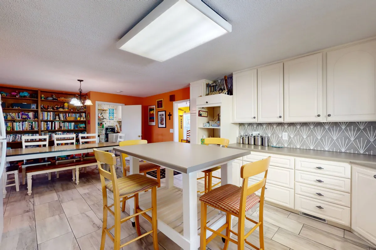 A cozy, inviting dining nook with a central table flanked by bookshelves, colorful wall art, and a kitchen area visible in the background. The space is designed for both communal meals and quiet reading, evoking nostalgia and the charm of a classic beach cottage.
