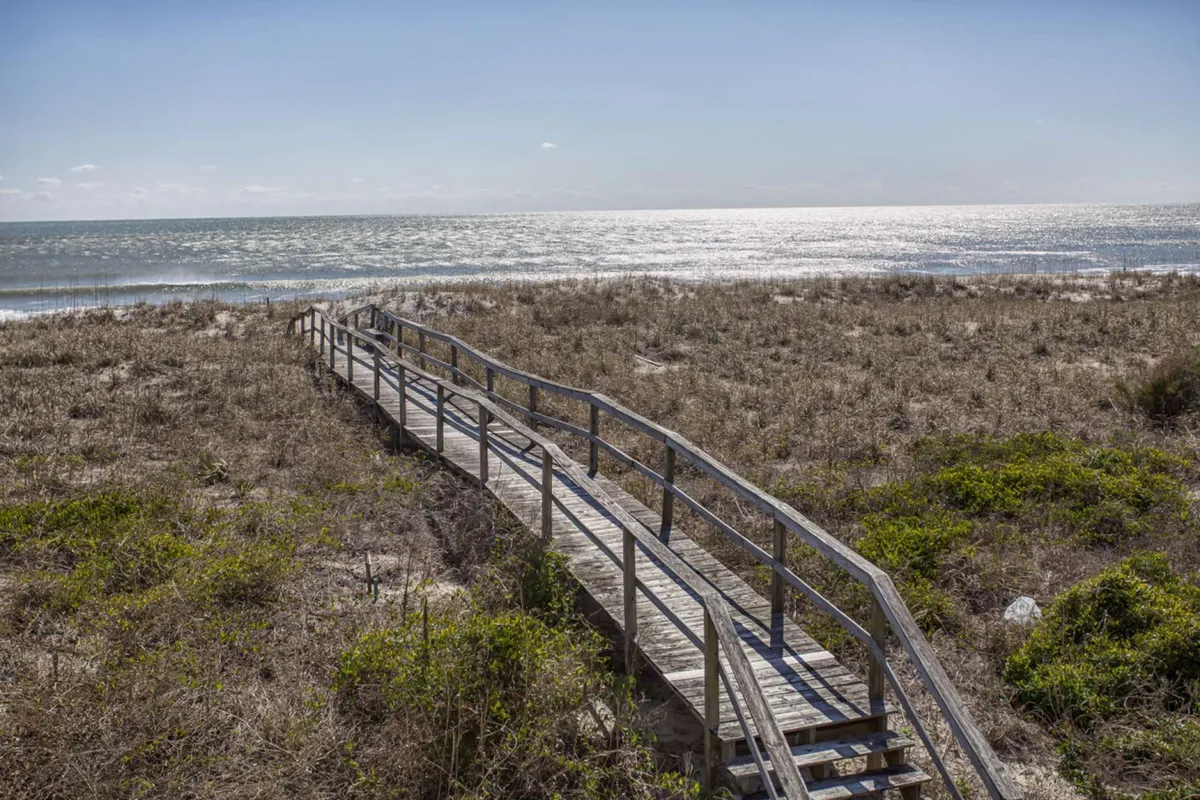 A tranquil scene featuring a wooden boardwalk winding through sand dunes, leading directly to the ocean. The soft light and gentle waves in the distance invite the viewer to imagine a peaceful stroll or a fun-filled beach day just steps from the property.