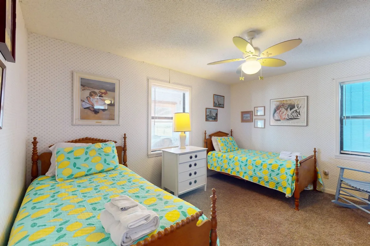 A cozy bedroom with two twin beds topped with colorful quilts, a white bedside table with lamp, and framed pictures on the wall. The atmosphere is cheerful and welcoming, ideal for guests seeking a shared yet private space.